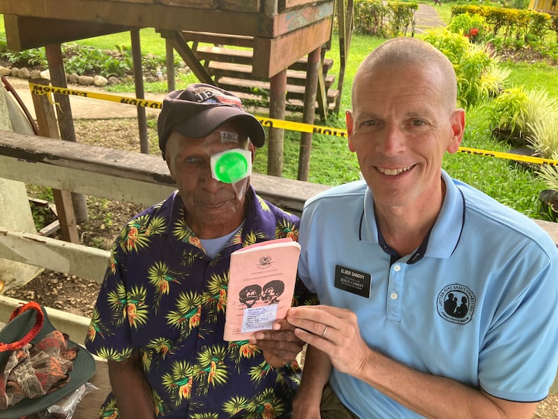 Pastor Ronny, left, takes a picture with Elder Bret Dandoy, right, while holding his medical record booklet after cataract surgery in 2026.