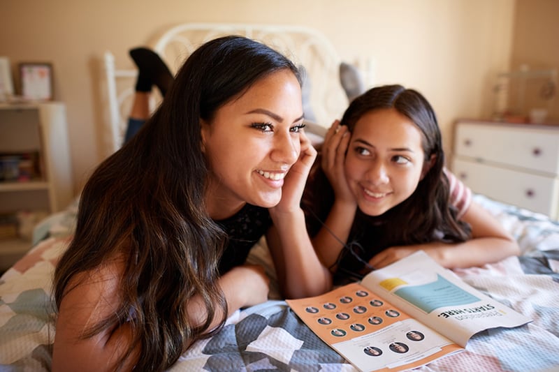 Two young girls lay on a bed looking at the General Conference Notebook together.