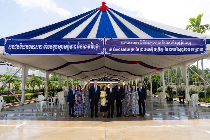 Elder Gerrit W. Gong and his wife, Sister Susan Gong, center, join four General Authority Seventies and their wives under a tent provided by the Cambodian government for the first day of Phnom Penh Cambodia Temple open-house tours on Wednesday, Aug. 12, 2026. The sign says: "Unity in religion brings peace and development. Congratulations on the Phnom Penh Cambodia Temple of The Church of Jesus Christ of Latter-day Saints."