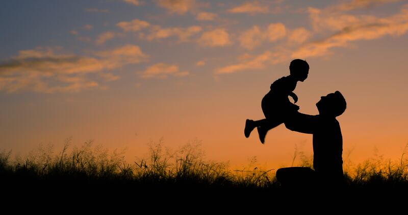 A kneeling father lifts up a child in a field against a setting sun.