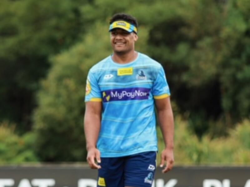 Joe Vuna smiles while walking across a rugby field. He wears a visor hat, cleats, a light blue shirt and dark blue shorts.