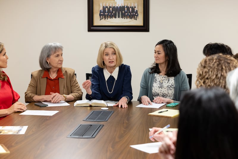 President Camille N. Johnson sits with her counselors at the head of a table and speaks to members of the Relief Society general advisory council.