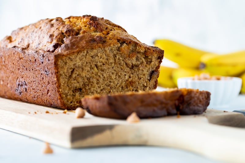 Straight on close up view of a banana bread with one slice in front, resting on a wooden board.