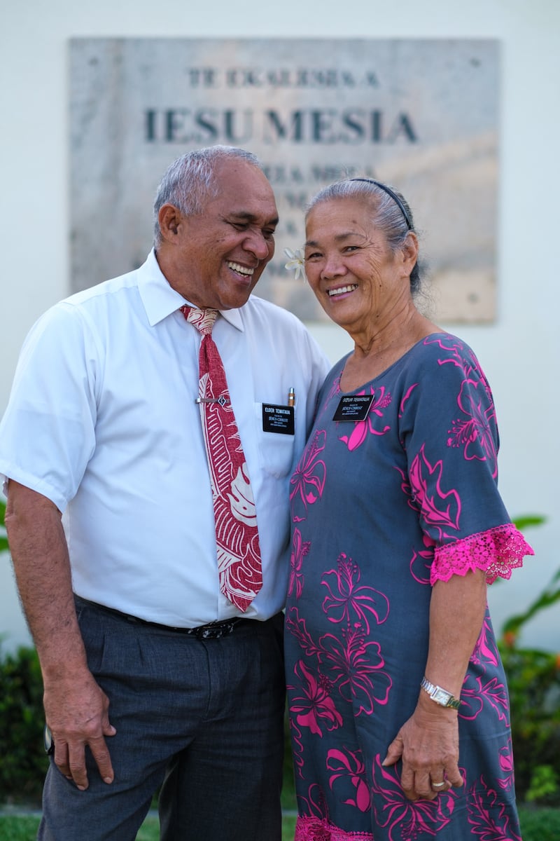 A senior missionary couple stands near a Latter-day Saint meetinghouse in Tahiti in 2026.