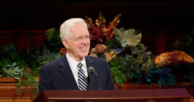 Man in a dark suit and glasses speaking at a wood podium with flowers behind him.