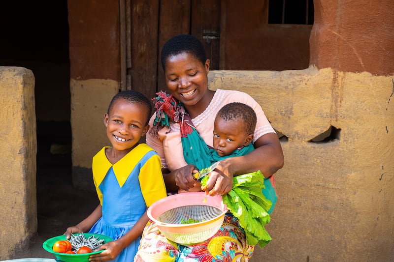 A young girl sits with her mother, who carries her 1-year-old sibling while chopping vegetables outside a house in southern Malawi.