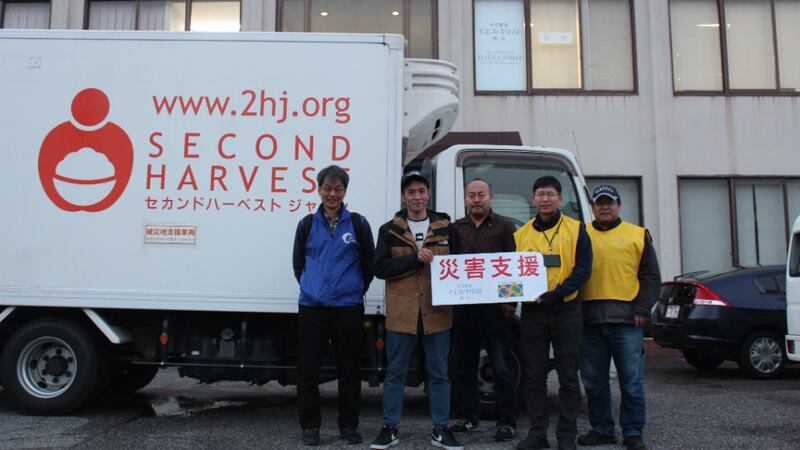 Two Church representatives in yellow Helping Hands vests present a donation to three men in front of a box truck with the words Second Harvest in English and Japanese.