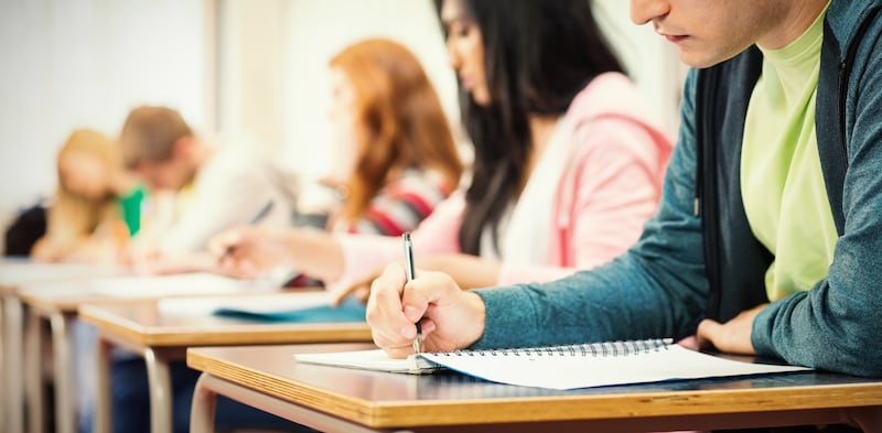 Students sit at a row of desks, writing in notebooks.