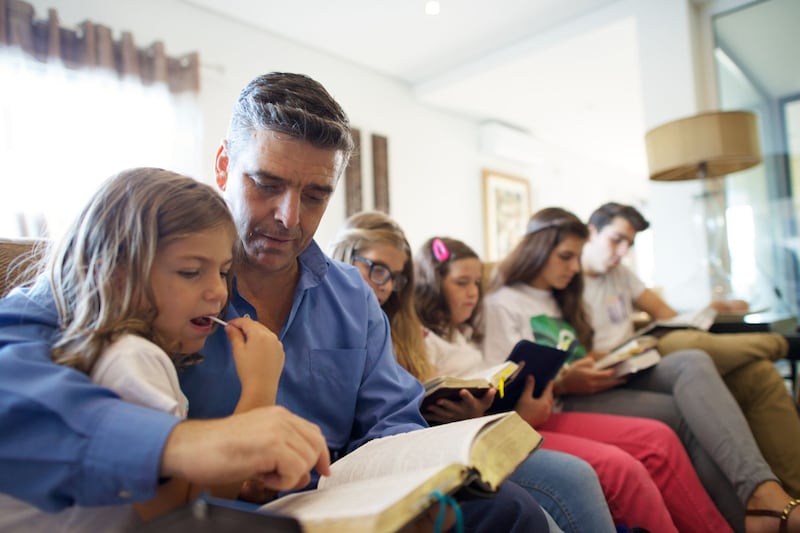 A father sits with his children in their living room to read from the scriptures together in Porugal.