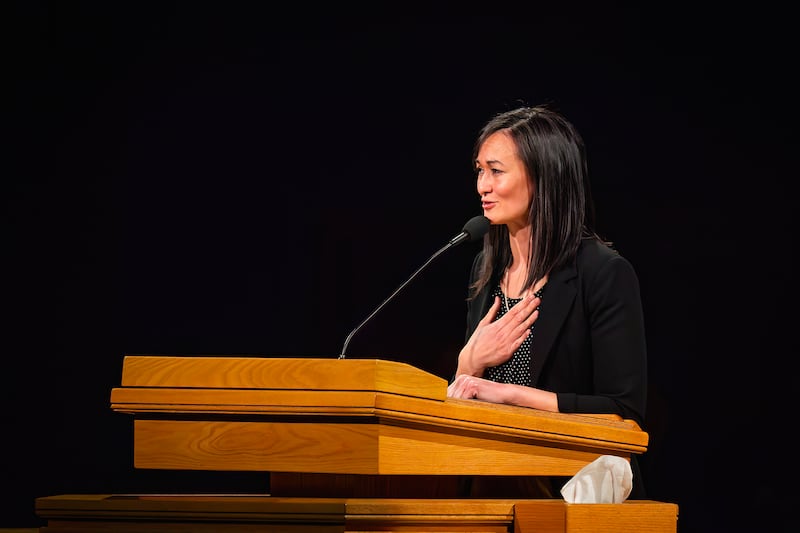Sister Yee, speaking at a wooden pulpit with a microphone, puts her right hand over her heart.
