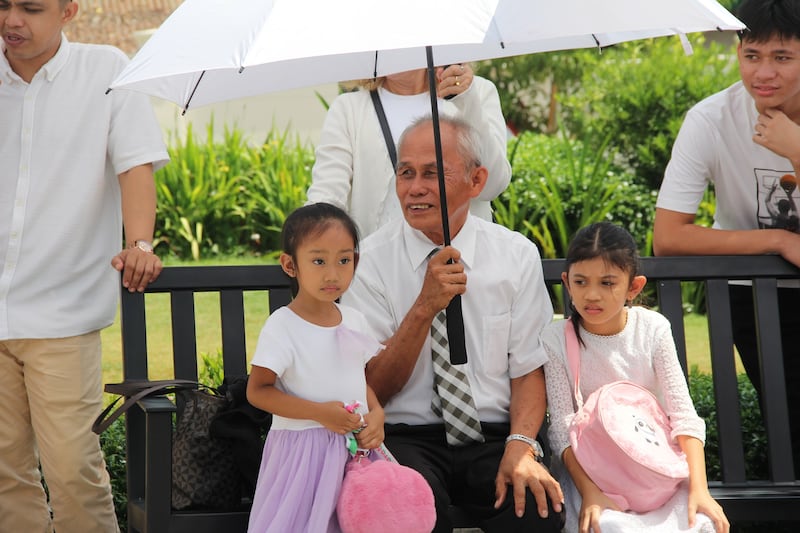 A grandfather sits under an umbrella with two girls on the grounds of the Davao Philippines Temple on the last day of the public open house, April 10, 2026.